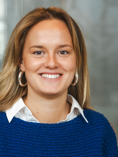 Person mit langen, hellen Haaren, blauem Pullover, weißem Hemd und runden Ohrringen, lächelt vor unscharfem Hintergrund.