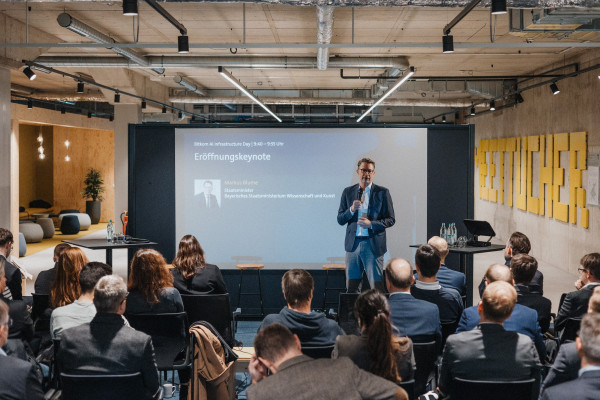 Markus Blume, Staatsminister, steht vor einer großen Leinwand, die seinen Speaker Slot ankündigt, mit Mikrofon in der Hand und spricht zu gut gefülltem Publikum auf dem AI Infrastructure Day.
