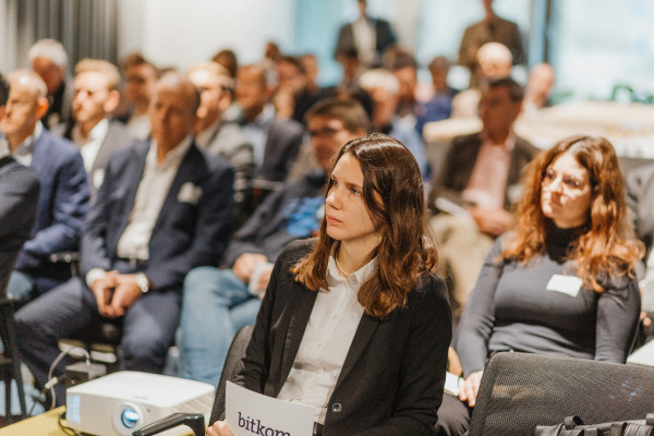 Im Bild ist das gut gefüllte Publikum des AI Infrastructure Day zu sehen, im Vordergrund eine Frau, dahinter mehrere weitere Reihen. Das Publikum hört aufmerksam zu, alle sind in Anzügen oder Business Attire gekleidet.