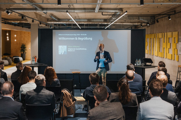 Niklas Veltkamp, Mitglied der Geschäftsleitung des Bitkom, steht vor einer großen Leinwand, die seinen Speaker Slot ankündigt, mit Mikrofon in der Hand und spricht zu gut gefülltem Publikum auf dem AI Infrastructure Day.