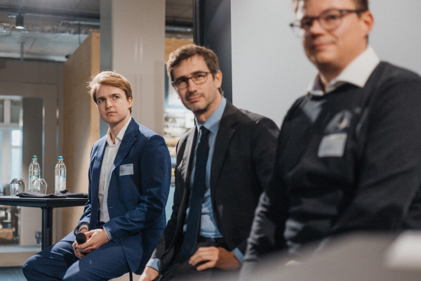 Drei Männer in Anzügen sitzen nebeneinander in einer Panel-Situation beim AI Infrastructure Day und hören der Moderatorin zu, die sich außerhalb des Bildes befindet.