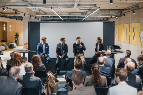 Drei Männer in Anzügen sitzen nebeneinander in einer Panel-Situation beim AI Infrastructure Day. Einer von ihnen spricht in ein Mikrofon, rechts daneben sitzt die Moderatorin. Im Vordergrund sind mehrere gut gefüllte Reihen Publikum zu sehen.