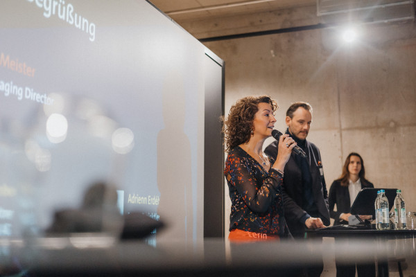 Eine Frau spricht vor einer großen Leinwand in ein Mikrofon, es handelt sich um einen Speaker Slot beim AI Infrastructure Day des Bitkom. Im Hintergrund des Bildes sind ein Mann und eine weitere Frau zu sehen.