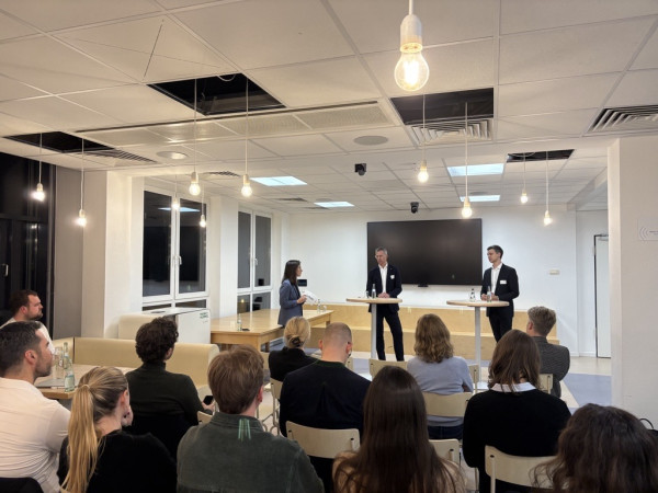 Drei Personen, eine Frau und zwei Männer, unterhalten sich an Stehtischen vor Publikum.