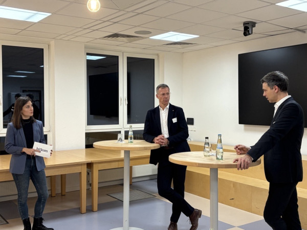 Drei Personen, eine Frau und zwei Männer, unterhalten sich an Stehtischen vor Publikum.