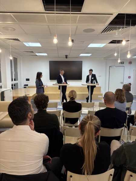 Drei Personen, eine Frau und zwei Männer, unterhalten sich an Stehtischen vor Publikum.