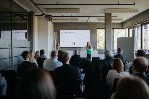 Konferenzraum mit großen Fenstern, zwei Frauen präsentieren, Text „PPWR - Was ist 2026 relevant?“ auf Leinwand.