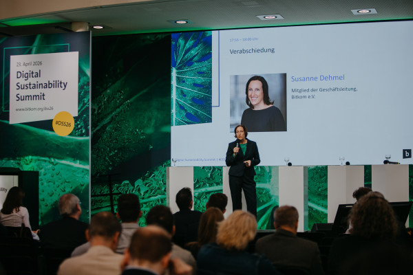 Frau mit dunklen Haaren spricht ins Mikrofon, Hintergrund zeigt Präsentation mit "Verabschiedung" und Namen. Publikum zuhört.