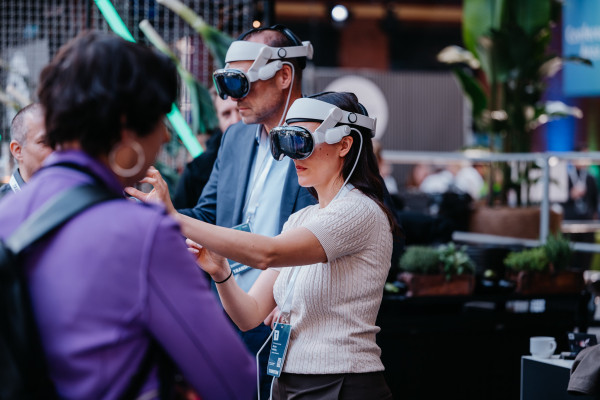 Mehrere Personen interagieren bei einer Veranstaltung, zwei tragen VR-Headsets, eine Frau gestikuliert.