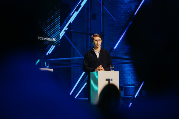 Mann am Podium mit Brille spricht, Glas Wasser auf dem Podium, geometrische Lichtinstallationen im Hintergrund.