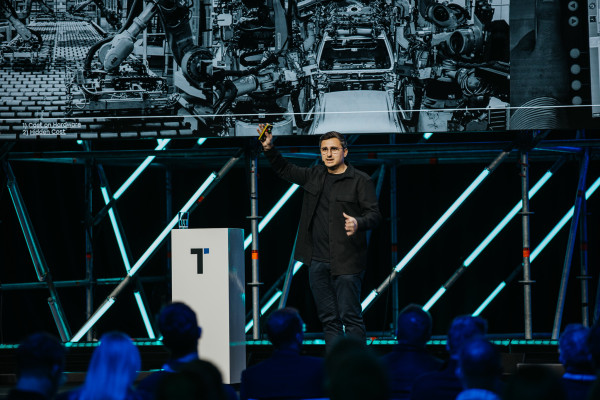 Person in black clothing gesticulating with a microphone on stage; technical display in background with listed points.