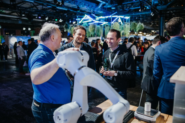 Mann mit grauen Haaren hält Roboterarm, umgeben von lächelnden Männern in modernem, beleuchtetem Raum.