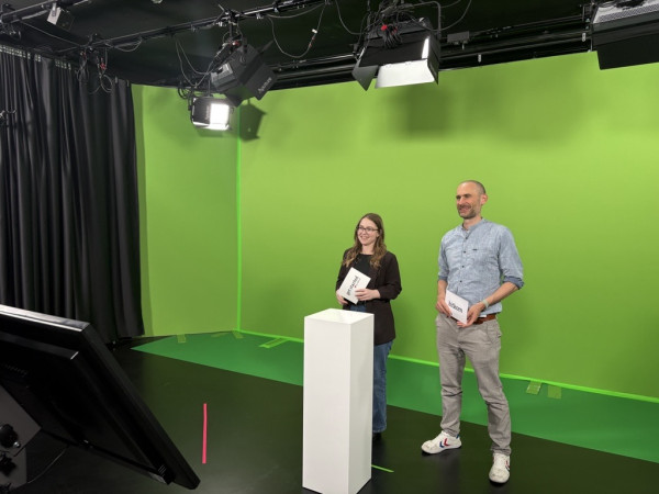 Zwei Personen in einem Studio mit grünem Hintergrund, beide halten Papier, vor einem weißen Podest und schwarzem Monitor.
