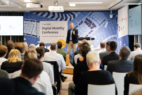 Redner auf Bühne spricht zu Publikum; Banner „7. Juli 2026 Digital Mobility Conference“ im Hintergrund.