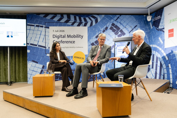 Drei Personen auf einer Bühne der Digital Mobility Conference 2026, zwei sitzen, eine spricht. Holztisch mit Wasser.