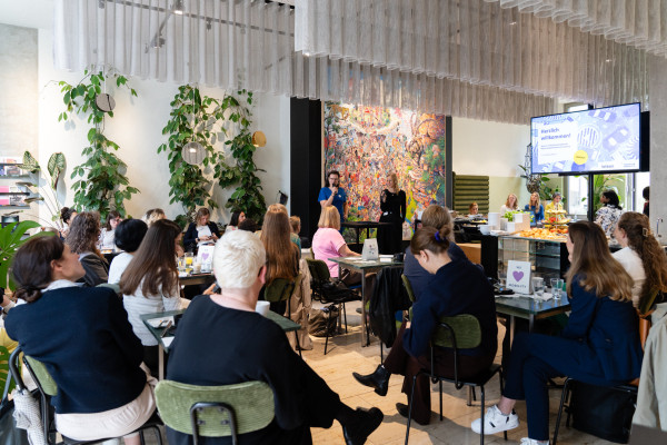 Konferenzraum mit sitzenden Teilnehmern, Präsentierenden, Snacks, Bildschirm mit „Herzlich willkommen!“ und Kunstwerk.