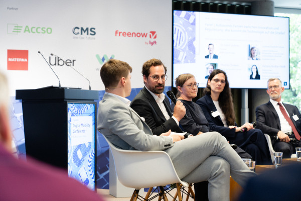 Mehrere Personen in formeller Kleidung diskutieren auf einer Bühne der Digital Mobility Conference. Rednerpult und Logos im Hintergrund.